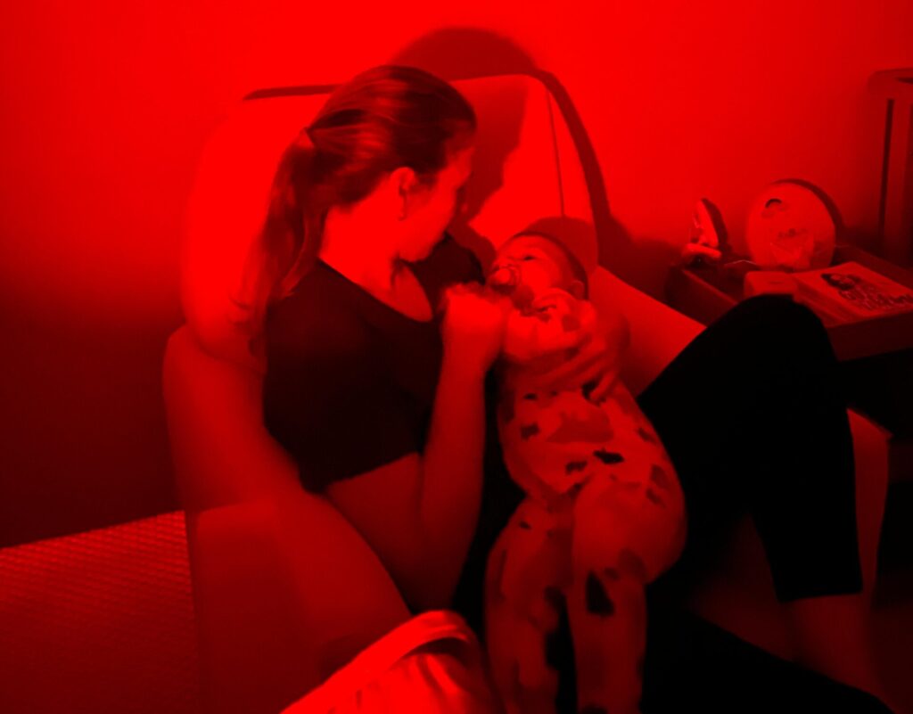 Mãe com bebê dormindo no colo em um quarto com luz vermelha, representando uma rotina de sono acolhedora.